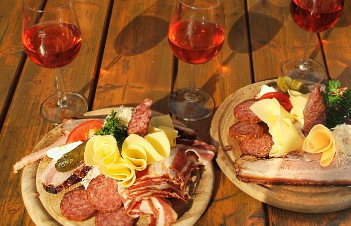 Två trätallrikar med olika sorters kallskuret, ostar, pickles och bröd står uppdukade på ett träbord tillsammans med tre glas rosévin. Solljuset framhäver den livfulla uppläggningen - perfekt efter en dag på skiferie i Østrig.