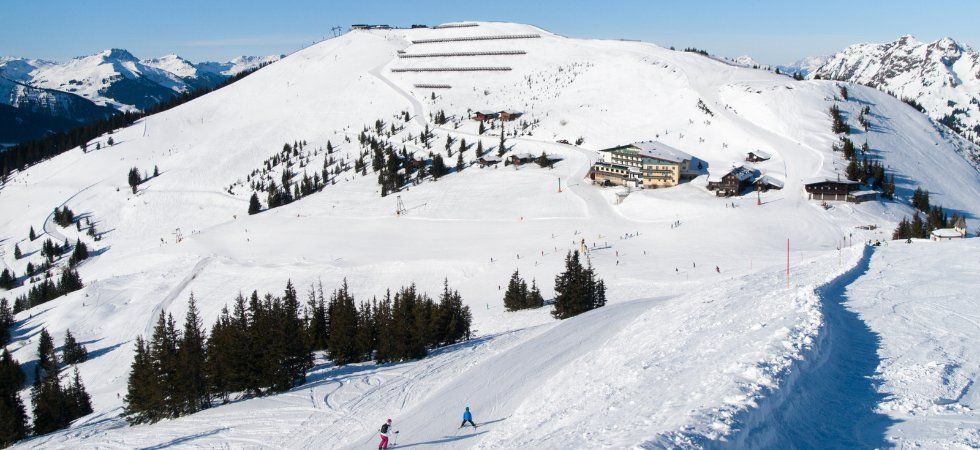 Ett snöigt berg med skidåkare i pisterna, tallar utspridda runt omkring och flera byggnader nära basen under en klarblå himmel - perfekt för en skiferie i Østrig.