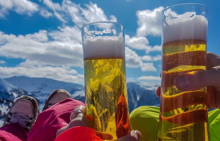 Två personer håller upp höga ölglas med skummande huvuden och njuter av en skiferie i Østrig utomhus, omgivna av snöiga berg och en ljusblå himmel med moln i bakgrunden.
