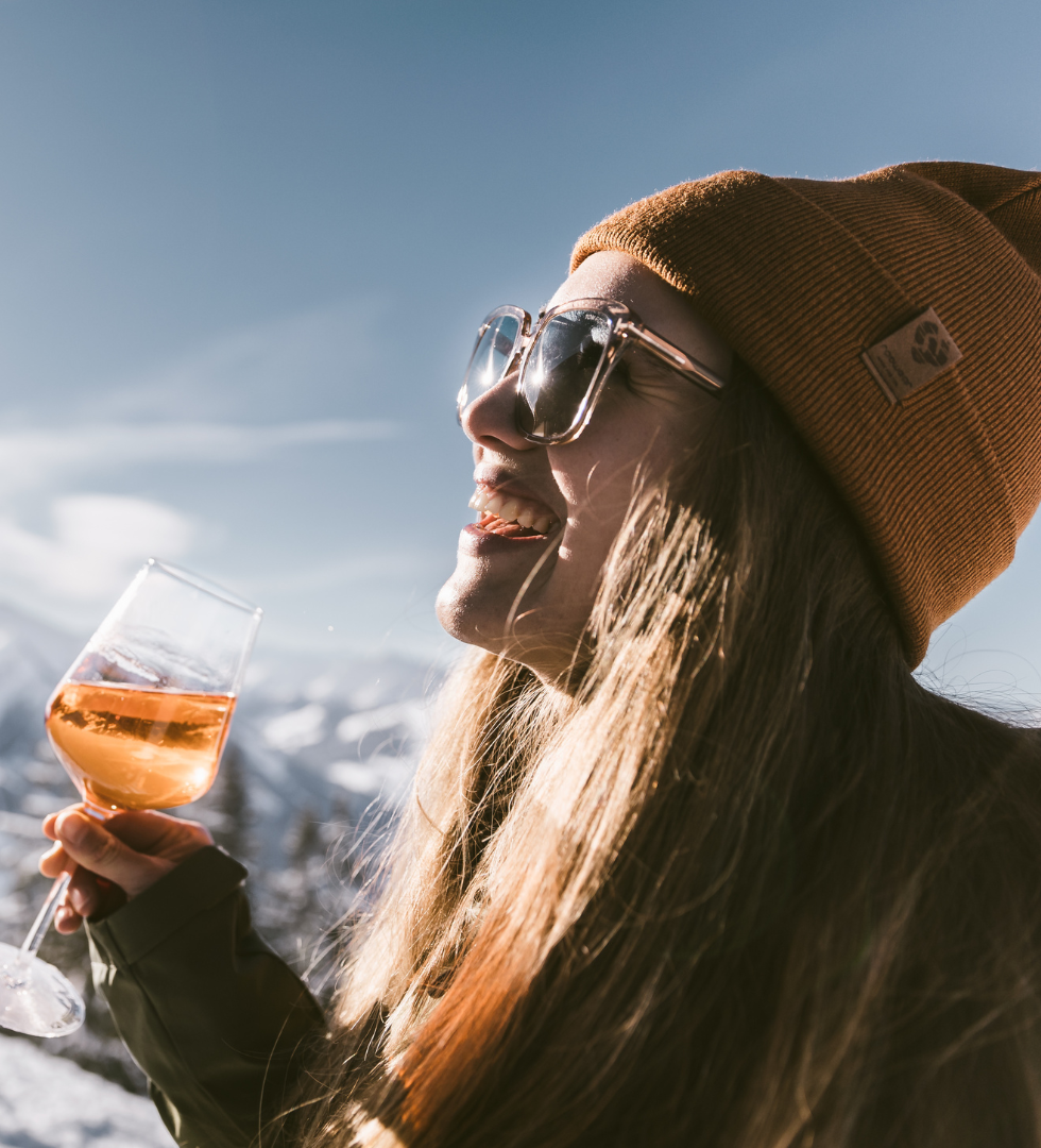 En person i en brun hue og solbriller smiler, mens han holder et glas vin udendørs. Baggrunden byder på snedækkede