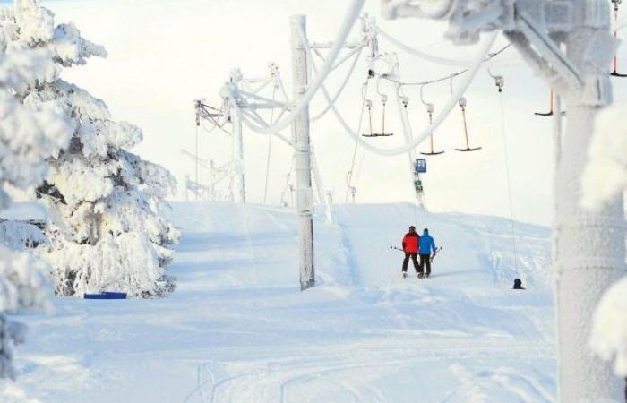 Två personer i vinterkläder står nära en skidlift på en snöig sluttning, omgivna av snötäckta träd och landskap under en klar himmel - perfekt för en skiferie i Sverige.