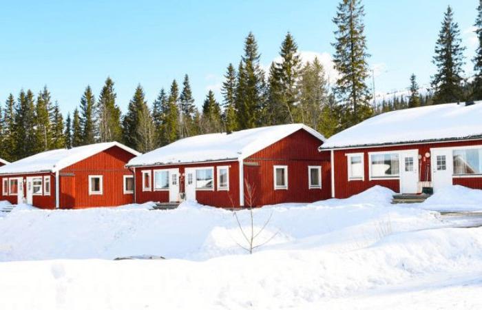 Fyra små röda trästugor med vit dekor ligger uppradade i ett snöigt landskap, omgivna av höga vintergröna träd under en ljusblå himmel - perfekt för en mysig skiferie i Sverige.
