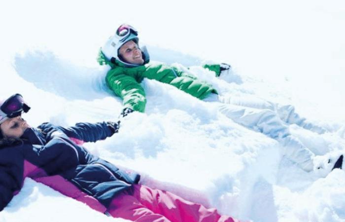 Två personer i vinterkläder ligger i snön och gör snöänglar, leende och avslappnade på sin skiferie i Sverige. Snön är ljus och tjock och varje person bär hjälm och glasögon.