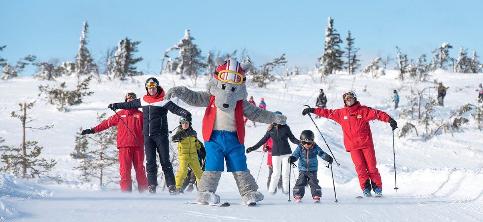 En person i en stor skidmaskotdräkt leder en grupp vuxna och barn som åker skidor i en snöig backe, omgiven av träd och klarblå himmel under deras skiferie i Sverige. Alla ser ut att njuta av vinteraktiviteten.