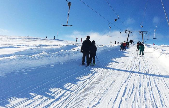 Människor njuter av en skiferie i Sverige, skidåkning uppför en snöig sluttning med hjälp av en skidlift en ljus, solig dag. Flera skidåkare prickar in backen under den klarblå himlen, deras skuggor sträcker sig över den glittrande snön.