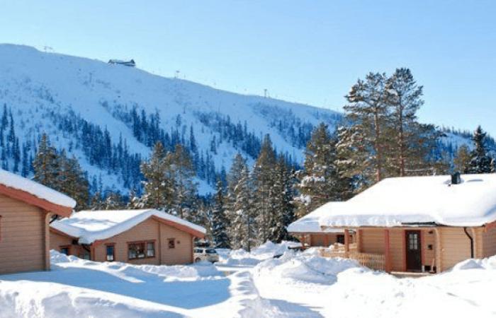 Snötäckta trästugor omges av höga tallar, med ett stort snötäckt berg i bakgrunden under en klarblå himmel - den perfekta miljön för ett skiferie i Sverige.