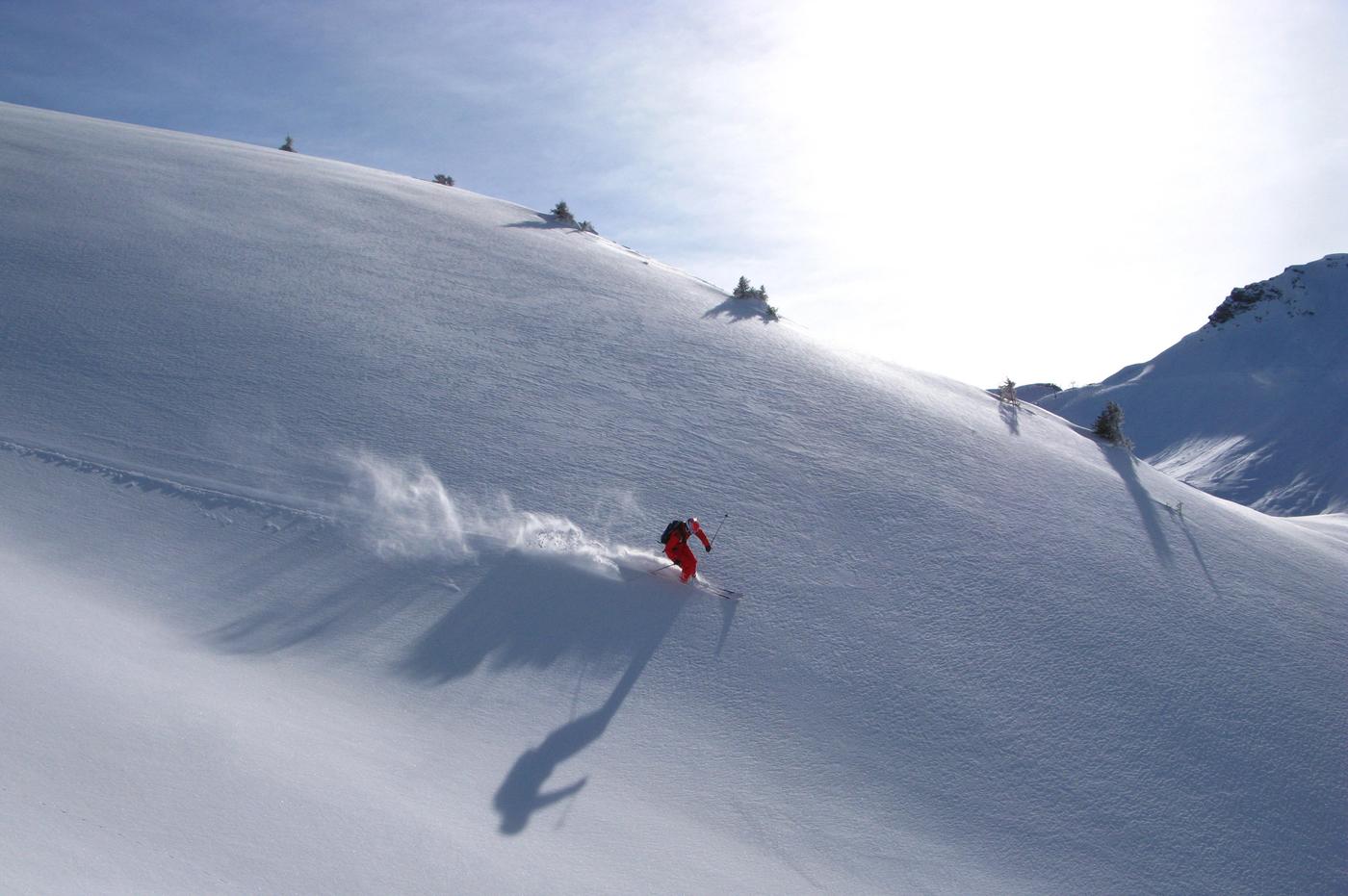 En skidåkare klädd i rött åker ner för en snöig bergssluttning i Westendorf och lämnar ett spår av puder efter sig. Solen lyser och kastar en lång skugga på den ostörda snön. Några små träd ligger utspridda längs åsen under en klarblå himmel.