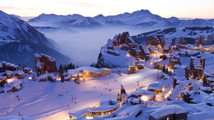 Snötäckt skidort i skymningen med upplysta byggnader inbäddade bland bergen i Westendorf. Landskapet inkluderar snöklädda toppar, dimma som rullar genom dalarna och en fridfull himmel som växlar från dag till natt, vilket skapar en pittoresk vinterscen perfekt för oförglömliga skidresor.