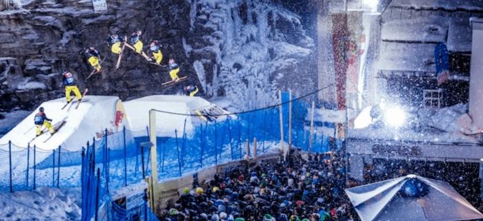 En skidåkare i gula byxor och blå jacka utför lufttrick från en snöramp på natten under en skiferie i Bad Gastein, med en jublande publik nedanför, blå skyddsnät, isiga klippor i bakgrunden och flera exponeringar som visar skidåkarens rörelse.