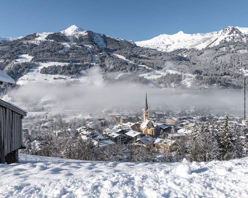 En snöig alpby med ett kyrktorn ligger nedanför skogsbeklädda berg under en klarblå himmel, med ett lager dimma som driver över staden.