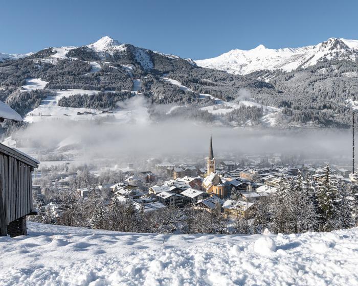 En snöig alpby med ett kyrktorn ligger nedanför skogsbeklädda berg under en klarblå himmel, med ett lager dimma som driver över staden.