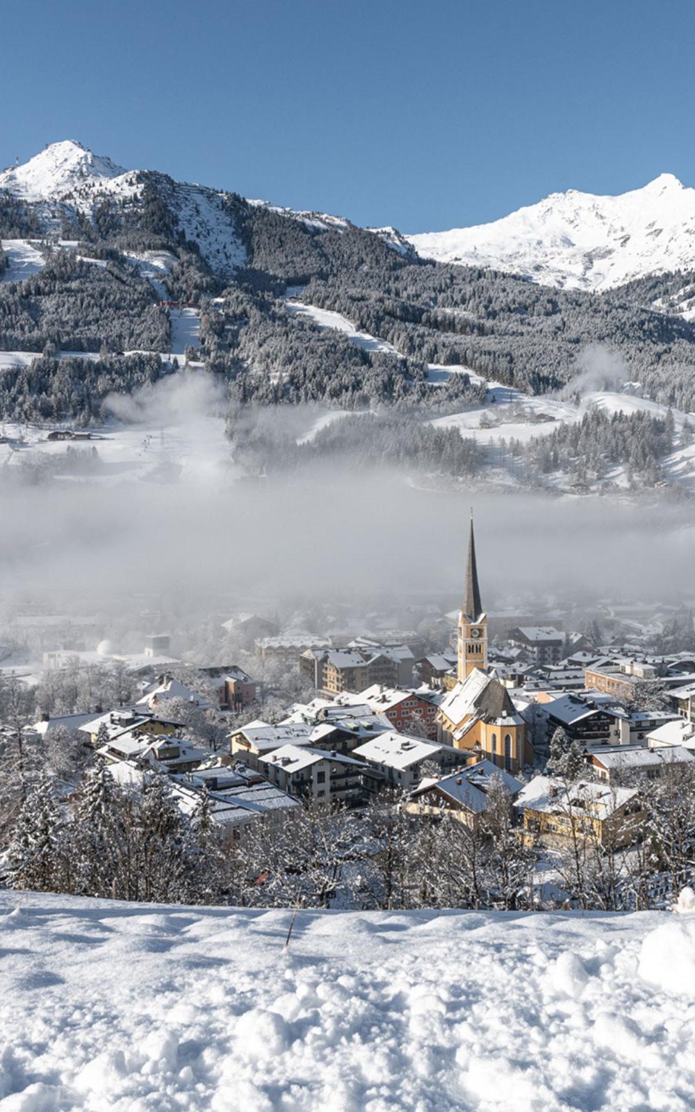Et vinterlandskab af en snedækket landsby med et fremtrædende kirketårn. Bjerge og skove, perfekt til skiferie, er dækket af sne under en klar blå himmel. Et lag lavthængende skyer lægger sig over landsbyen og skaber en fredfyldt atmosfære ideel for skirejser.