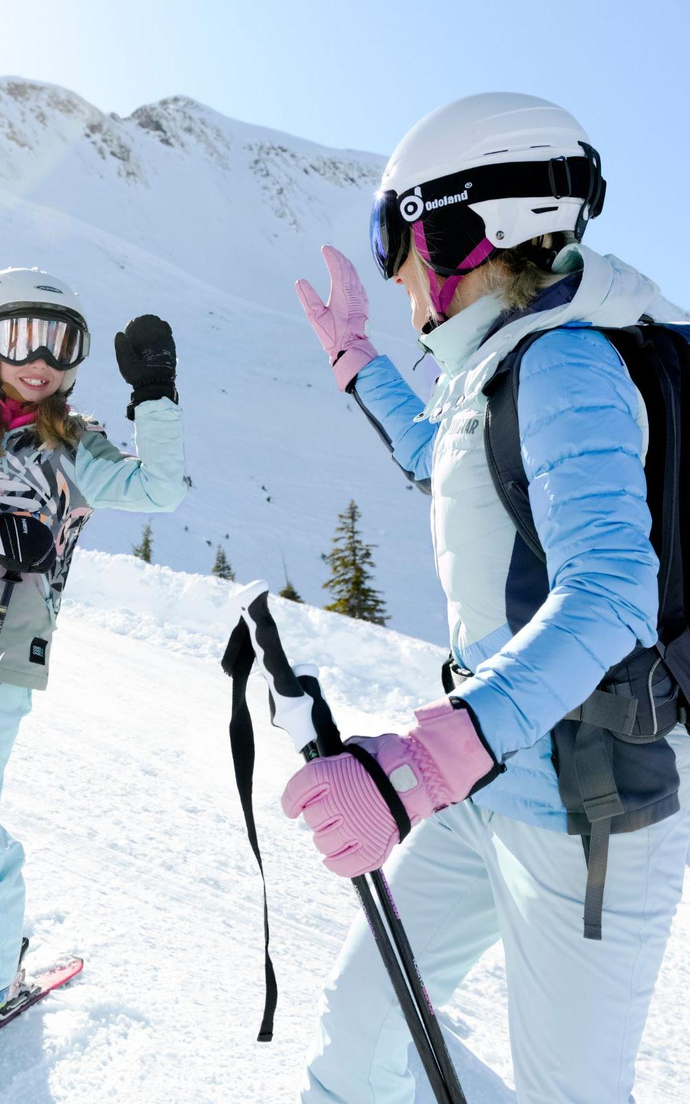 To skiløbere, iført hjelme og briller, deler en high-five på en sneklædt skråning under deres skiferie. Den ene er en voksen med rygsæk og skistave, den anden et barn. De griner midt i Westendorfs solbeskinnede bjerge under en klar blå himmel.