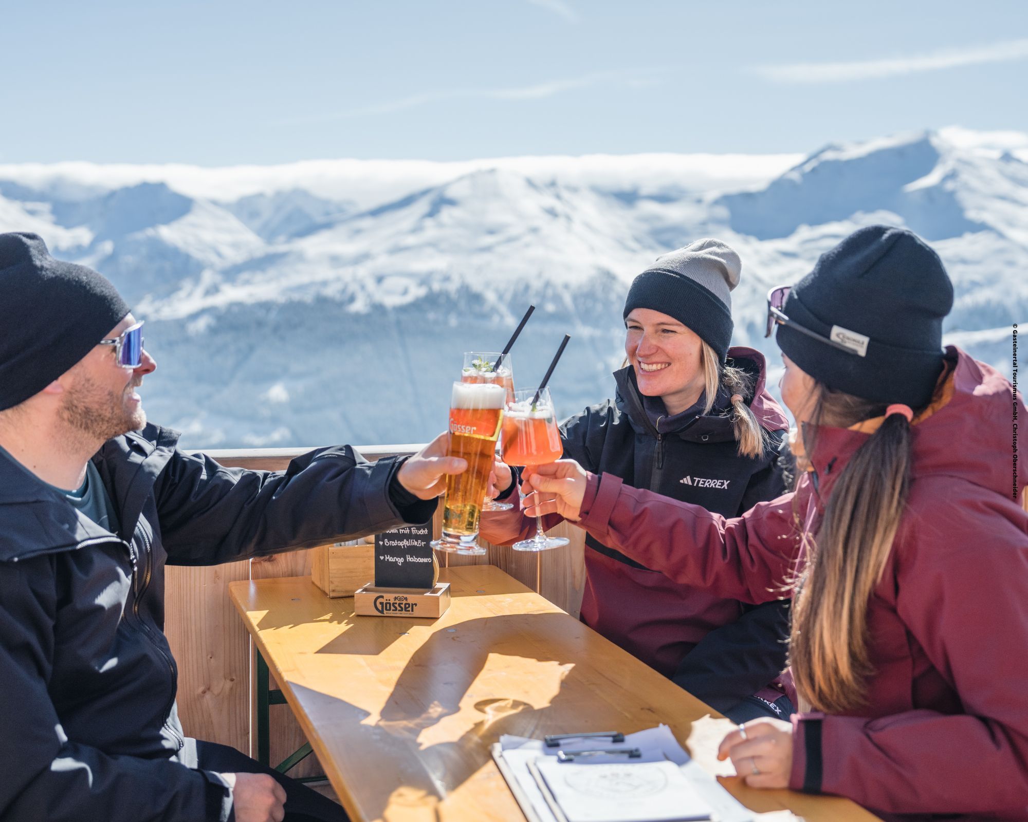Tre personer i vintertøj klirrer i glas ved et udendørs bord mod de sneklædte bjerge i Wagrain. De smiler og nyder drinks med skarpt sollys, der fremhæver scenen, og fanger et perfekt øjeblik af glæde midt i Østrigs fantastiske vinterlandskab.