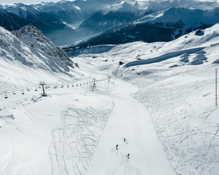 Flygfoto över en snöig skidbacke med tre skidåkare på väg ner, omgiven av snötäckta bergstoppar och en skidlift till vänster. Spåren går kors och tvärs över snön under en molnig, solbelyst himmel.