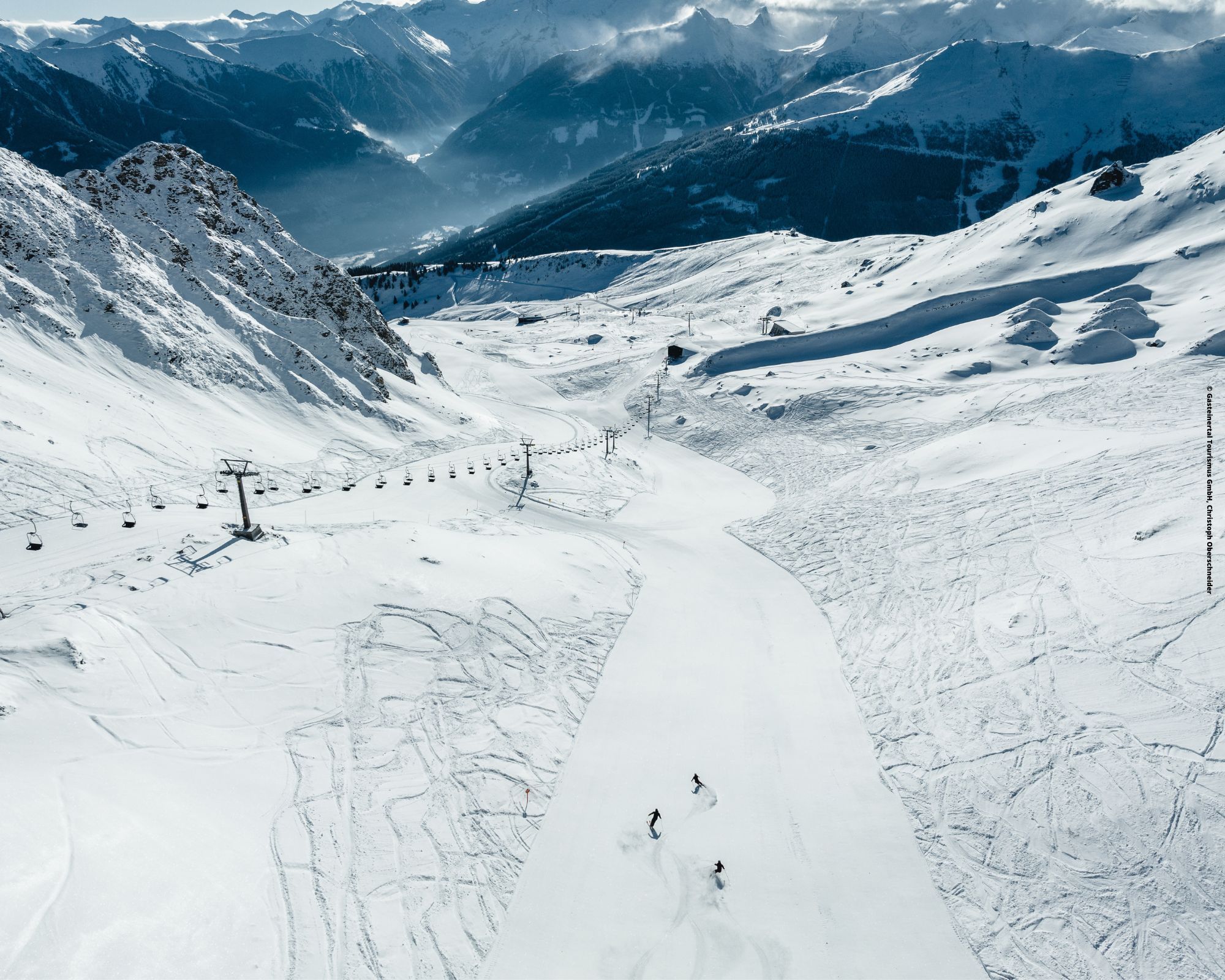 Luftfoto af et snedækket bjerglandskab i Saalbach med to skiløbere på en bred skråning. Snedækkede tinder og træer langs scenen, med en fjern bjergkæde under en delvist overskyet himmel perfekt til en skiferie. En skilift løber parallelt med pisten til venstre.