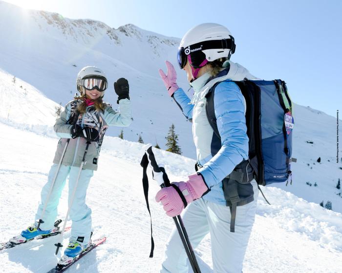 Två personer iförda skidutrustning och hjälm gör high five med varandra på en snöig bergssluttning, med skidstavar i handen och starkt solljus som lyser upp det snöiga landskapet.