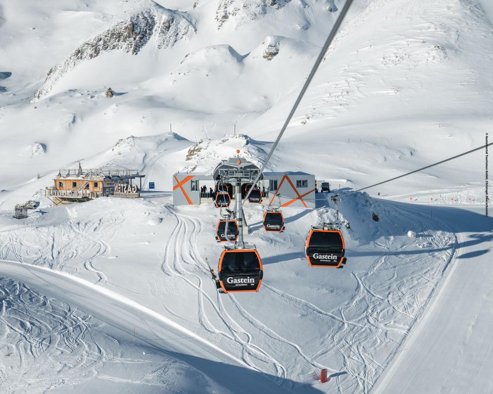 Flera orange och svarta gondolbanor märkta Gastein färdas uppför en snöig bergssluttning mot en skidliftstation, med skidspår synliga i den vita snön och bergen i bakgrunden.