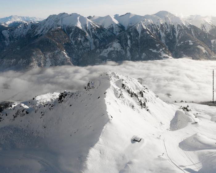 En sneklædt bjergtop rejser sig storslået med fjerne sneklædte områder i baggrunden. Et tæppe af skyer dækker dalen og skjuler delvist dette fantastiske Saalbach-landskab. Sollys kaster skygger hen over sneen og fremhæver det barske terræn - en perfekt ramme om et skiferieeventyr.