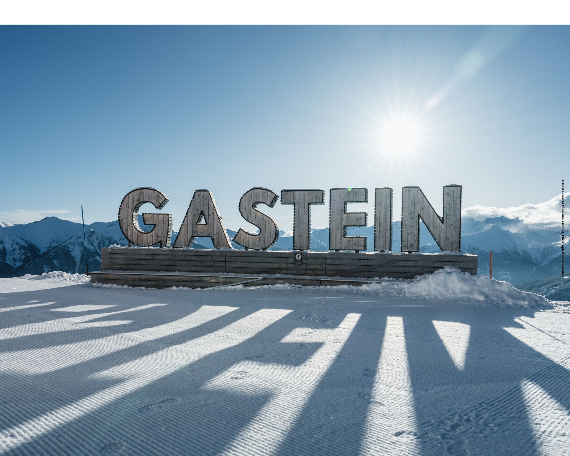 Et stort skilt, der staver GASTEIN står på en snedækket platform under en klar himmel. Det er en indbydende scene for skirejseentusiaster, mens solen skinner klart over fjerne bjerge og kaster lange skygger på den snedækkede jord, der minder om Saalbachs fortryllende vinterlandskaber.
