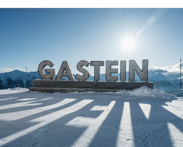 Stora metallbokstäver med bokstäverna GASTEIN står på en snöig plattform med berg i bakgrunden. Solen skiner starkt bakom skylten och kastar långa skuggor på snön.