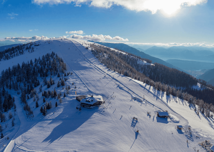 Flygfoto över en snötäckt bergssluttning med skidspår, spridda träd och två byggnader; perfekt för skidresor i Wagrain när solen skiner över avlägsna berg under en delvis molnig himmel.