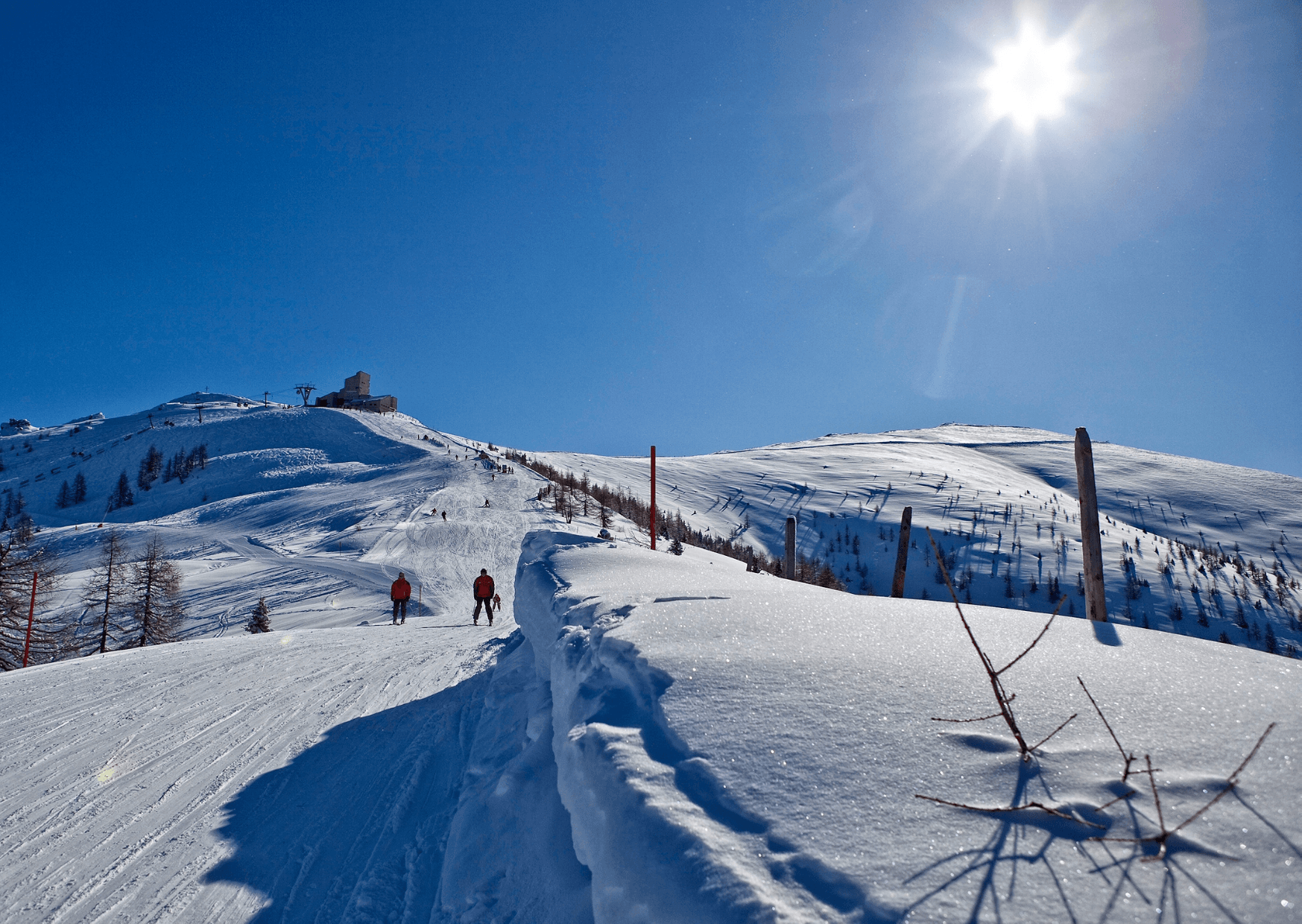 En solrig vinterscene i Westendorf med to personer, der går op ad en sneklædt bakke. Himlen er klar, og solen skinner klart. I baggrunden kan en bygning og træer ses på en fjern skråning, hvilket fuldender denne fantastiske udsigt dækket af nysne.