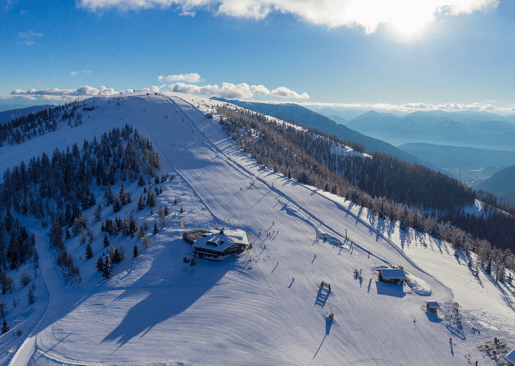 En sneklædt bjergskråning under en klar blå himmel med spredte skyer sætter scenen for en mindeværdig skiferie. I Saalbach går skiløjper på kryds og tværs af bakken, mens en lille bygning ligger nær centrum. Solen skinner klart i det fjerne og kaster sit lys over dette maleriske landskab.
