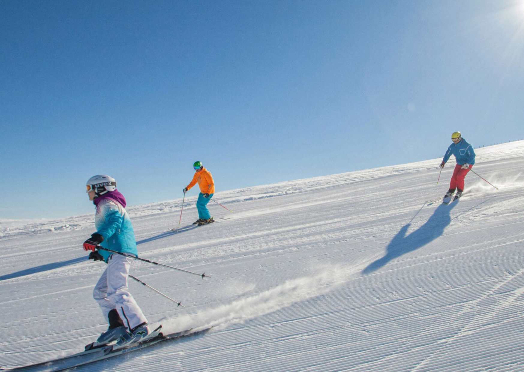 Tre personer i färgglada skidkläder njuter av en skidresa, åker skidor nerför en ljus, solig backe i Saalbach och lämnar spår i den nypreparerade snön under en klarblå himmel.