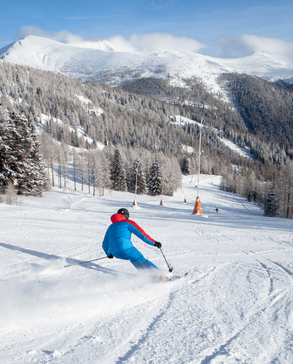 En person i blåt og rødt skitøj står på ski ned ad en sneklædt skråning i Wagrain, omgivet af snedækkede træer og bjerge i baggrunden under en klar blå himmel.