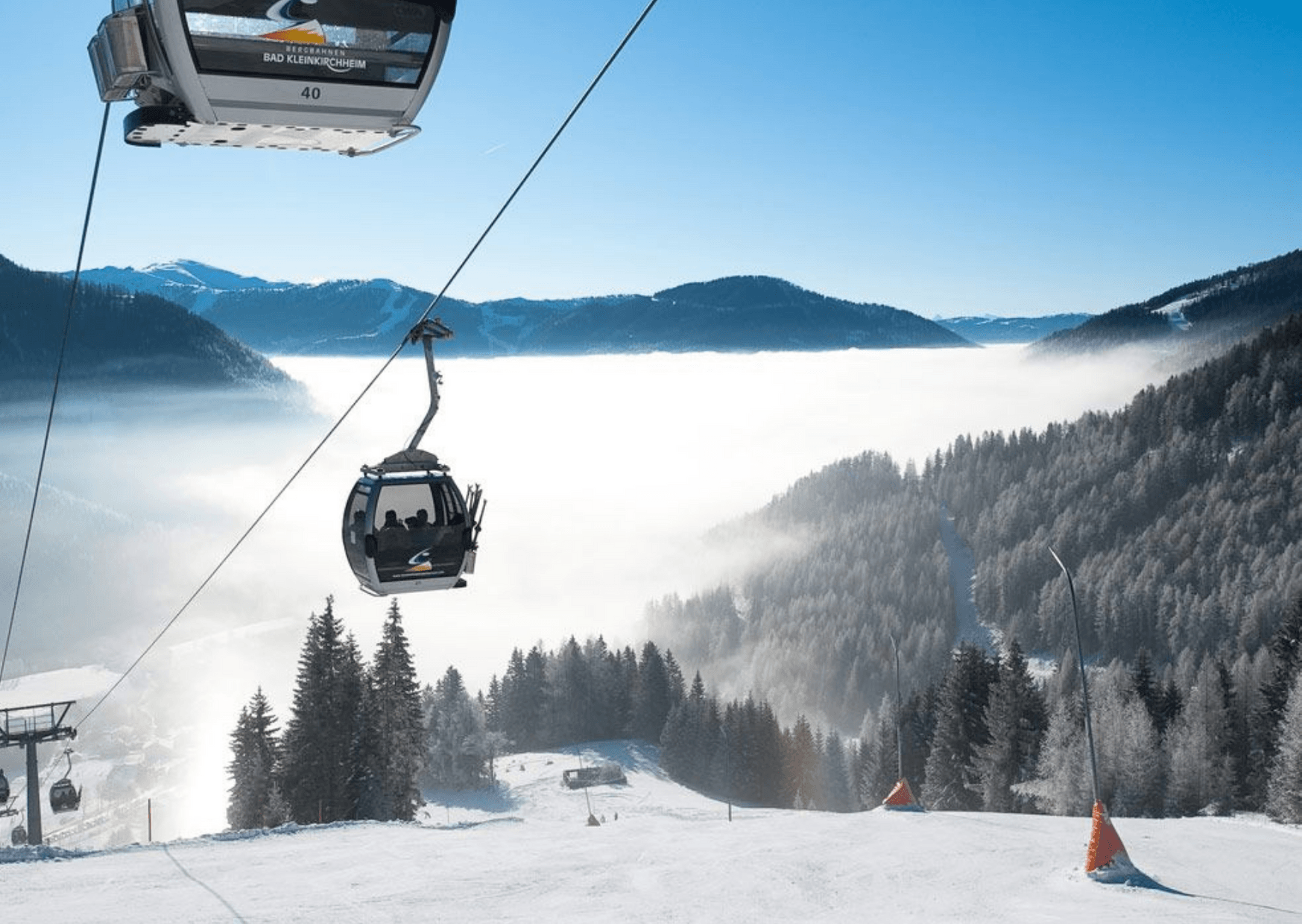 Skidliftstolar stiger upp över en snötäckt bergssluttning i Wagrain, med utsikt över tallar och en dimmig dal, med majestätiska berg och blå himmel i bakgrunden – perfekt för ett skidåkningsäventyr.