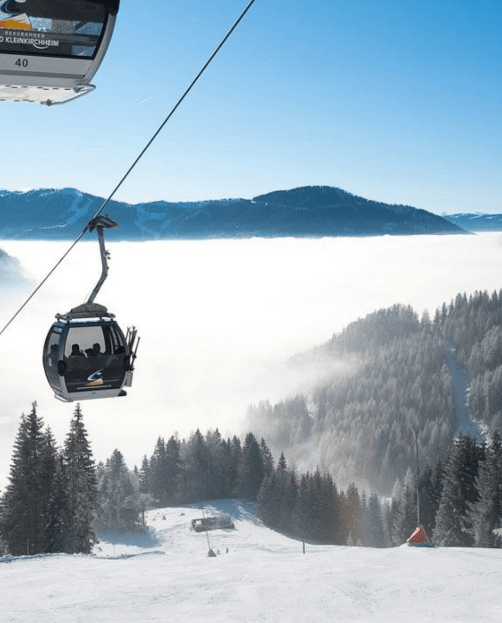 En naturskøn udsigt over et sneklædt bjerglandskab i Wagrain med stolelifte, der glider over en skibakke. Solen skinner klart, og et tågelag svæver over dalen nedenfor, omgivet af skovklædte bakker og fjerne tinder under en klar blå himmel. Perfekt til spændende skirejseeventyr.