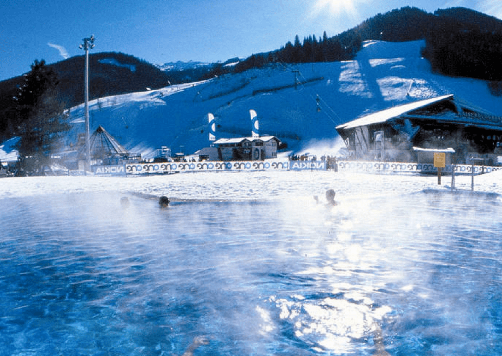 I Saalbach simmar folk i en ångande utomhuspool omgiven av snö och skidbackar, med synliga berg och skidliftar under en klarblå himmel – en perfekt miljö för en minnesvärd skidresa.