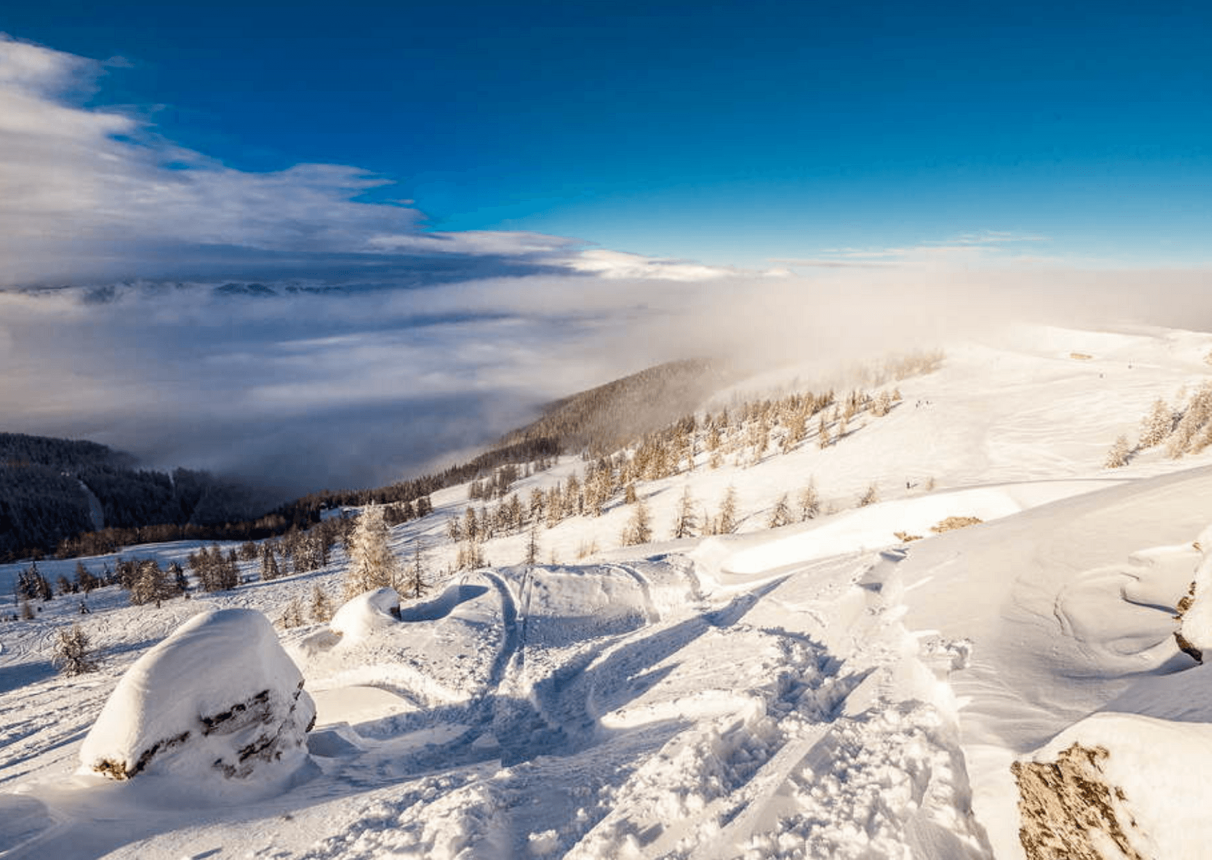 Et betagende vinterlandskab i Westendorf viser snedækkede bakker og træer under en klar blå himmel. Bløde skyer svæver over en fjern bjergkæde og tilføjer dybde til den fredfyldte scene, perfekt til skirejser. Skygger skaber tekstur på det snedækkede terræn.
