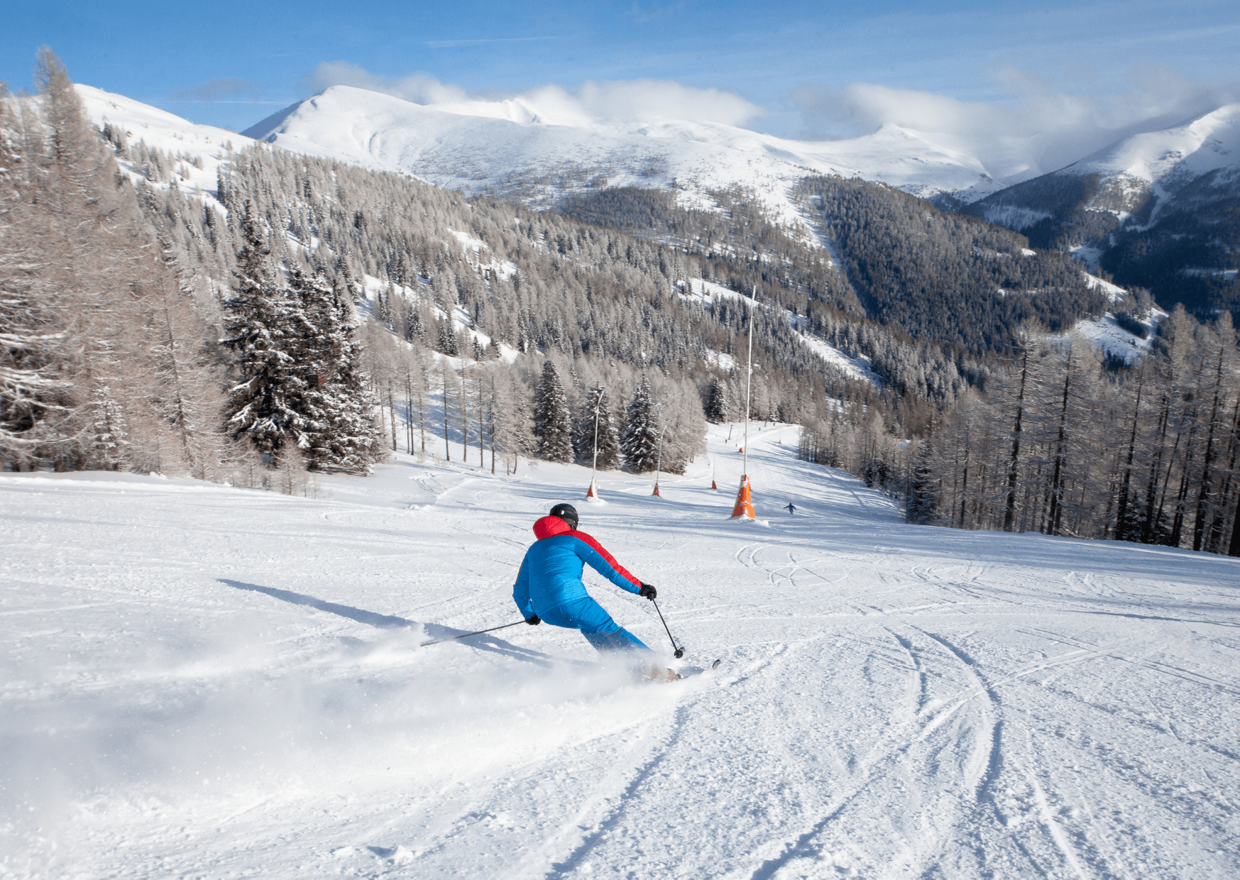 En skidåkare i blå och röd dräkt glider nerför en snöig bergssluttning i Saalbach, omgiven av snötäckta träd och avlägsna berg under en delvis molnig himmel.