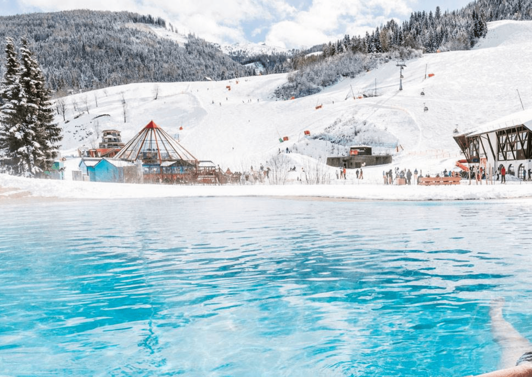 En klarblå utomhuspool ligger i förgrunden av en snötäckt skidort i Saalbach, med skidåkare, tallar och byggnader i stugstil synliga under en delvis molnig himmel – perfekt för oförglömliga skidresor.