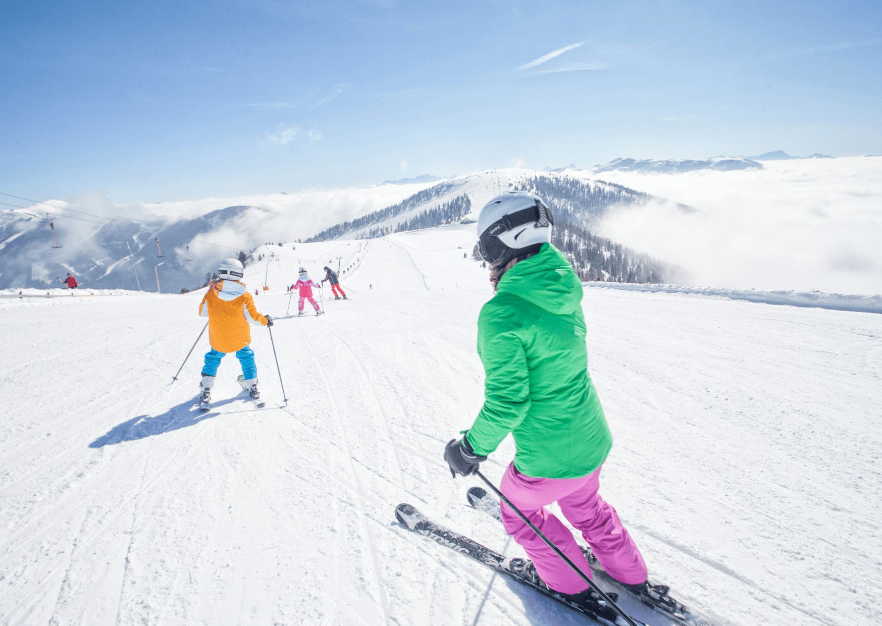 Två personer i färgglada skidjackor och hjälmar åker skidor nerför en snöig bergssluttning i Westendorf en solig dag, med avlägsna skidåkare och snötäckta toppar synliga under den blå himlen – perfekt för oförglömliga skidresor.