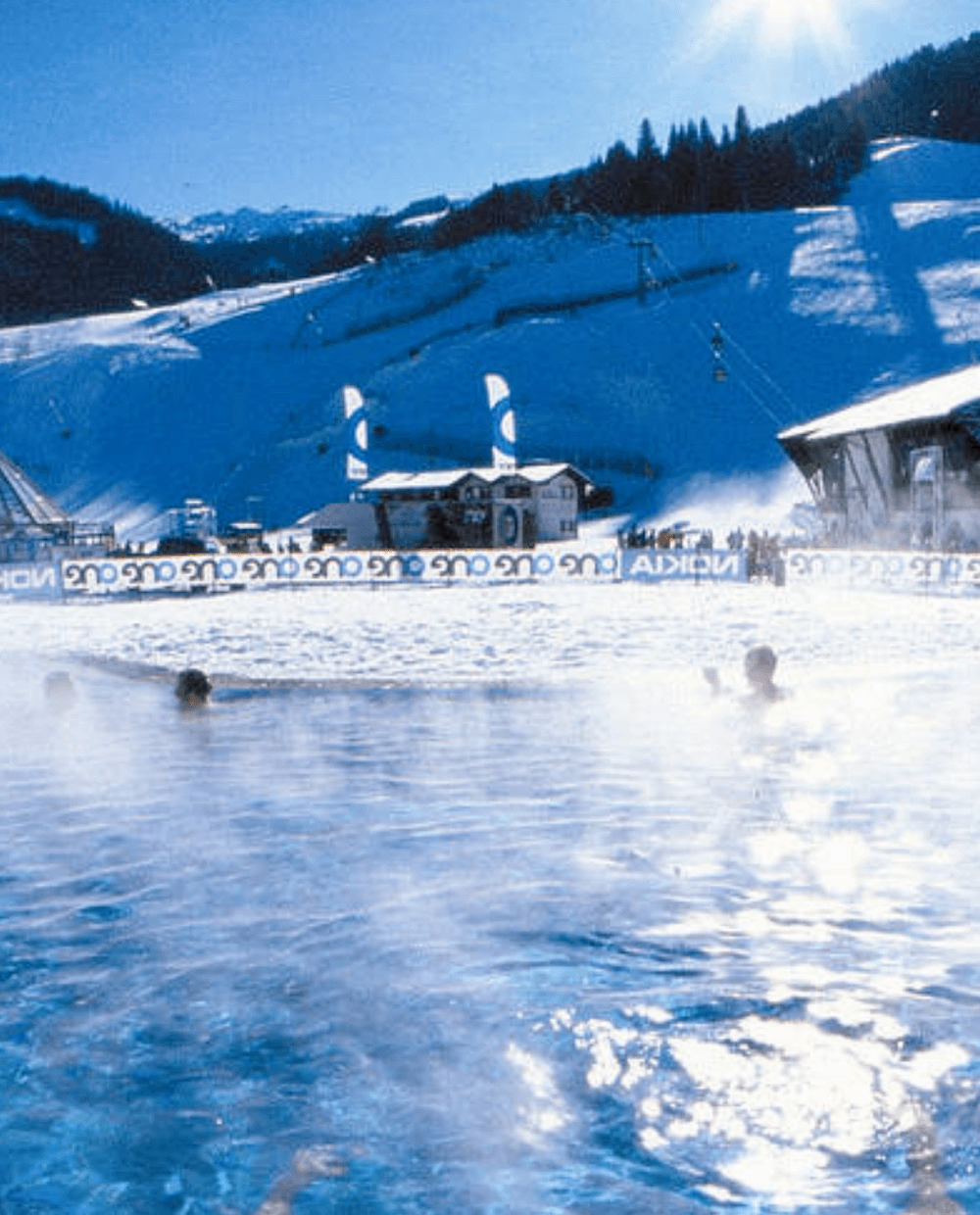 En dampende udendørs pool med mennesker, der svømmer på baggrund af sneklædte bjerge. Skilifter og en lodge er synlige i det fjerne under en klar blå himmel, og fanger den perfekte essens af en skiferie i Saalbach eller Wagrain.