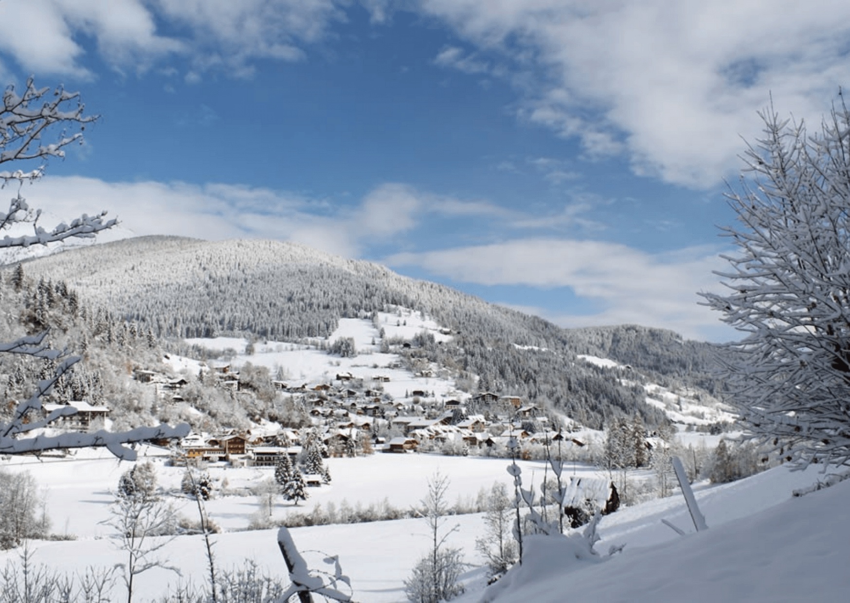 En malerisk snedækket landsby beliggende i en dal, der minder om Westendorf, med huse spredt ud over landskabet. Sneklædte træer og bølgende bakker omgiver området under en delvist overskyet blå himmel.