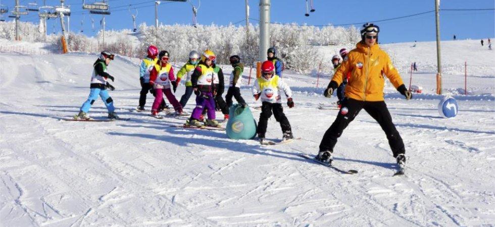 En skidlärare i orange jacka leder en grupp barn på skidor nedför en snöig backe under en klarblå himmel under en skiferie i Beitostølen, med skidliftar och snötäckta träd i bakgrunden.