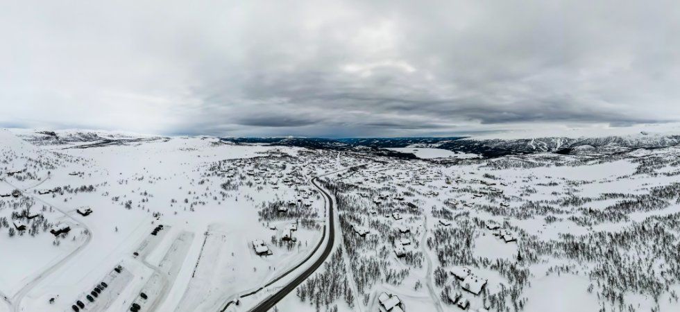 En flygbild med panoramautsikt över ett snöigt landskap med utspridda hus, slingrande vägar och berg i fjärran under en molnig himmel - perfekt för ett skiferie i Beitostølen.