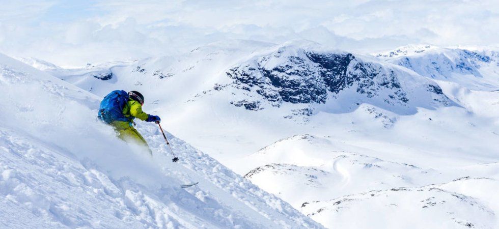 En skidåkare i ljusa kläder njuter av en skiferie i Beitostølen och glider nerför en snöig bergssluttning omgiven av snötäckta toppar under en molnig himmel.