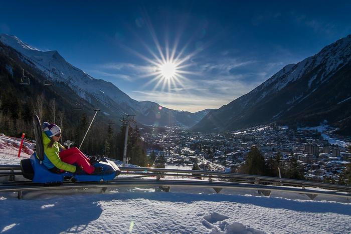En person åker berg-och-dalbana på snötäckta stigar i Saalbach, omgiven av snötäckta berg, under en strålande sol med strålar som lyser över en pittoresk dalstad nedanför – perfekt för oförglömliga skidåkningsäventyr.