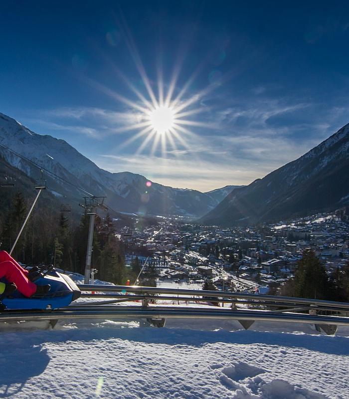 En person åker berg-och-dalbana på snötäckta stigar i Saalbach, omgiven av snötäckta berg, under en strålande sol med strålar som lyser över en pittoresk dalstad nedanför – perfekt för oförglömliga skidåkningsäventyr.