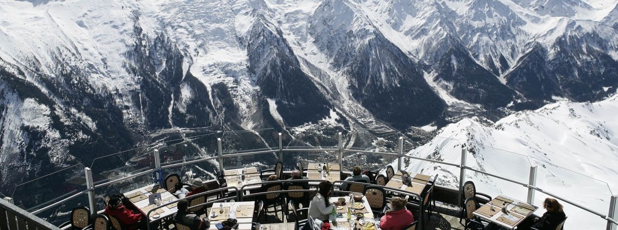 En gruppe mennesker spiser på en udendørs terrasserestaurant, der ligger på et bjerg i Westendorf. Snedækkede tinder og en dal giver en fantastisk kulisse, med klar himmel, der forstærker den naturskønne skiferieudsigt. Der er dækket borde til spisning, omgivet af panoramisk skønhed.