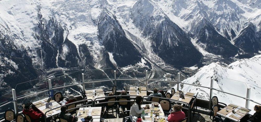 En gruppe mennesker spiser på en udendørs terrasserestaurant, der ligger på et bjerg i Westendorf. Snedækkede tinder og en dal giver en fantastisk kulisse, med klar himmel, der forstærker den naturskønne skiferieudsigt. Der er dækket borde til spisning, omgivet af panoramisk skønhed.
