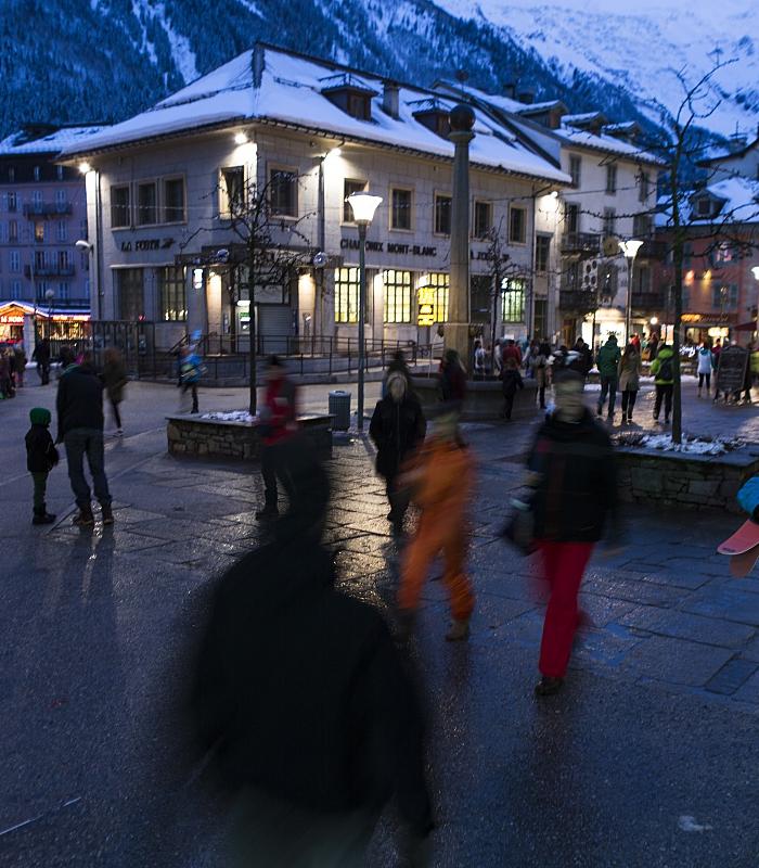 Människor går genom ett livligt torg i Westendorf i skymningen, omgivet av snötäckta byggnader och berg; vissa bär skidutrustning, och färgglada vinterkläder sticker ut mot kvällens skidåkningsscen.