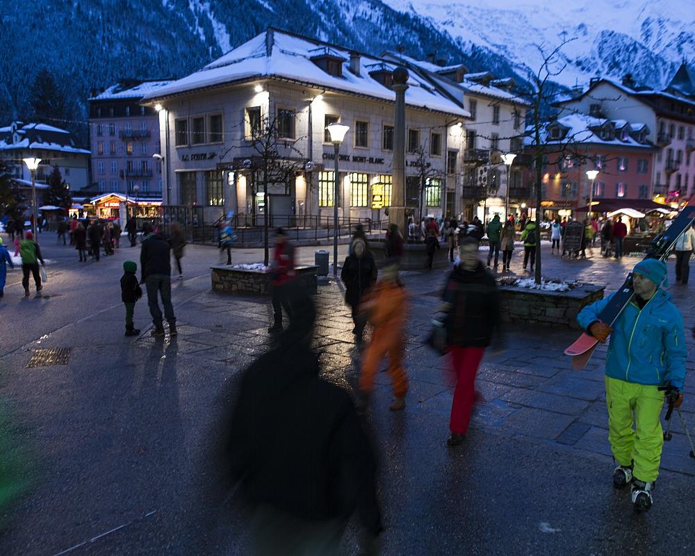 Människor går genom ett livligt torg i Westendorf i skymningen, omgivet av snötäckta byggnader och berg; vissa bär skidutrustning, och färgglada vinterkläder sticker ut mot kvällens skidåkningsscen.