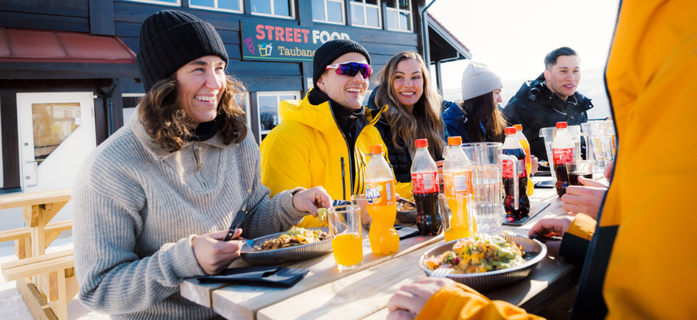 En grupp vinterklädda människor sitter vid ett bord utomhus och njuter av mat och dryck under sin skiferie i Geilo, de ler och skrattar tillsammans, med en byggnad märkt Street Food i bakgrunden.