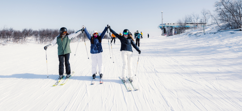 Tre skidåkare i vinterkläder håller varandra i handen och höjer armarna medan de står i en snöig skidbacke under en Skiferie i Geilo, med träd och en skidlift i bakgrunden under en klar himmel.